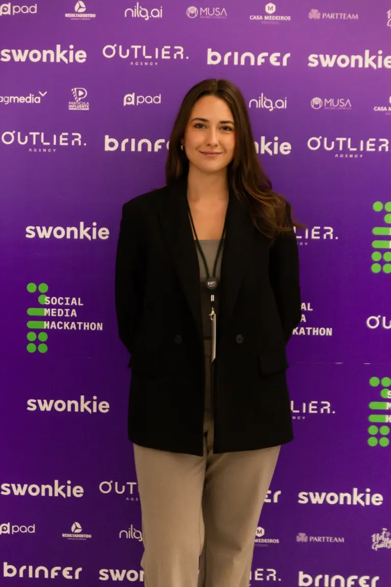 Mulher com cabelo castanho, vestida com terno preto e calça bege, posando diante de um fundo roxo com vários logotipos e...