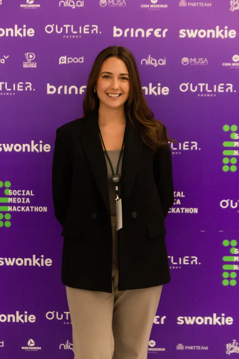 Mulher sorridente vestindo um blazer preto, camisa cinza e calça bege, em evento com fundo roxo com várias logos e nomes...