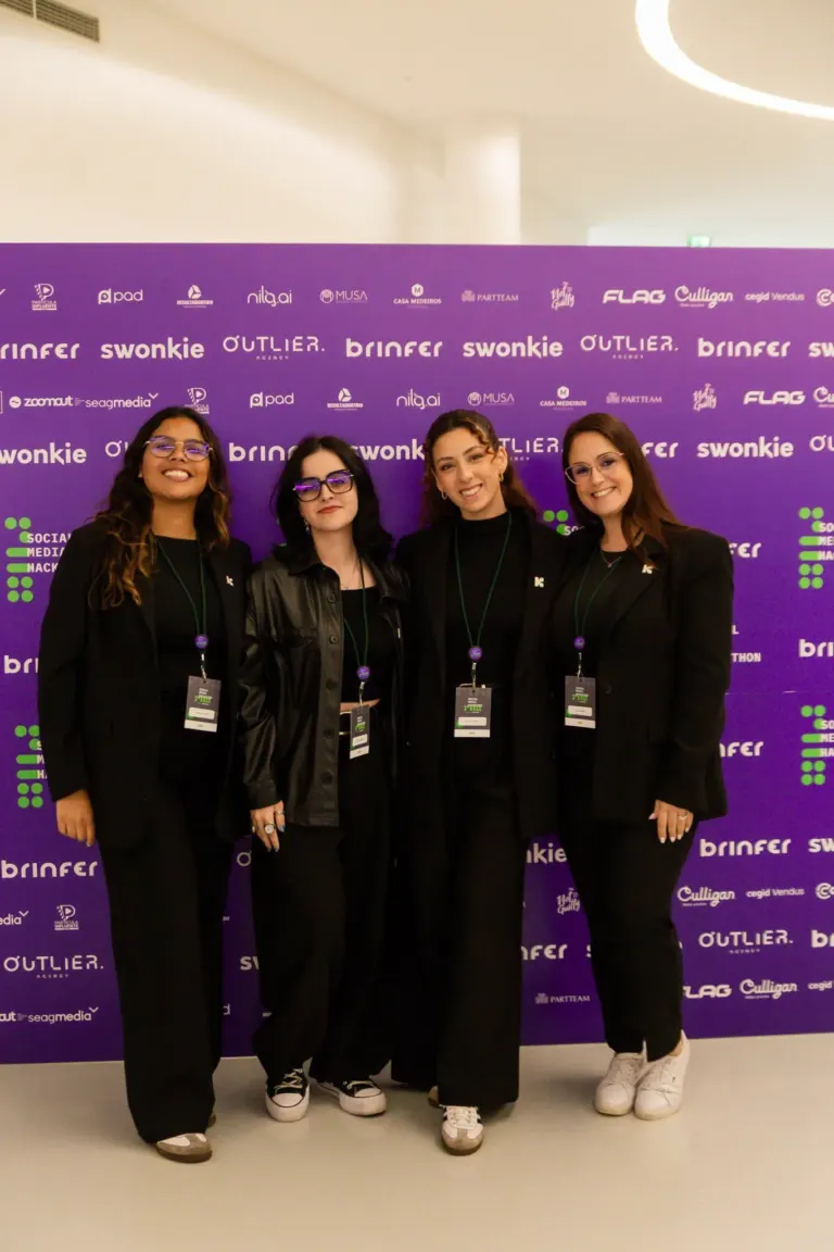 Quatro mulheres sorridentes usando roupas pretas e crachá, posando juntas em frente a uma parede roxa com logos de...