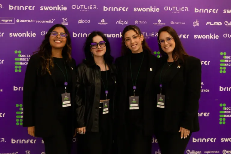 Quatro mulheres sorrindo e posando juntas em frente a um painel roxo com logotipos de patrocinadores e o título 'Social...