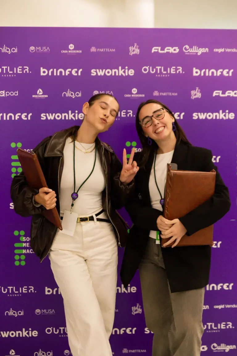 Duas mulheres posando juntas ao fundo de uma parede roxa com logos de marcas e eventos, uma delas faz sinal de paz com a...