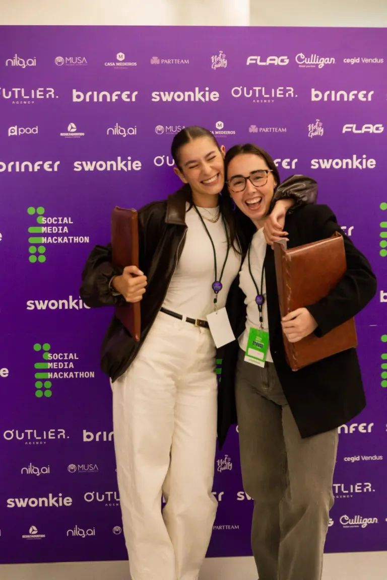 Duas mulheres sorrindo e abraçando na frente de um fundo roxo com logos e texto relacionados a eventos de tecnologia e...