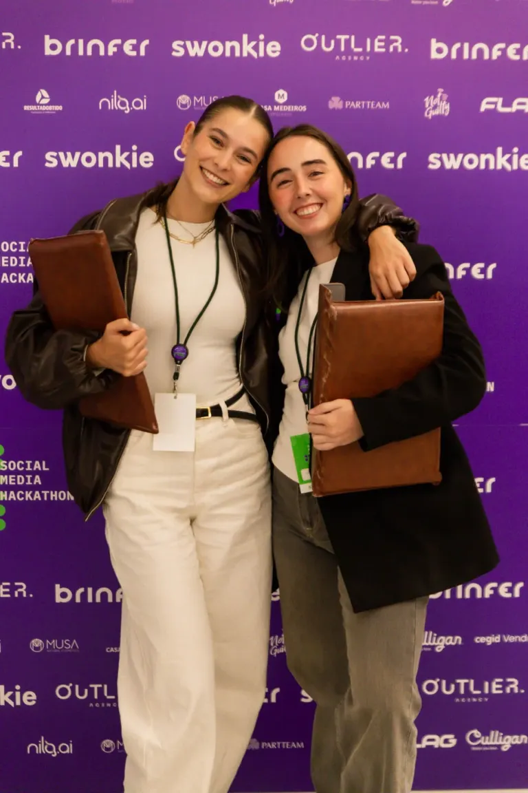 Duas mulheres sorrindo, abraçadas, segurando pastas, posando diante de um fundo roxo com logos de eventos e marcas.