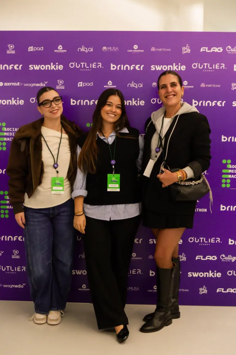Três mulheres sorrindo e posando juntas contra um fundo roxo com logos de empresas e marcas.