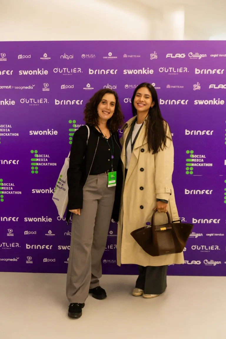 Duas mulheres sorrindo posando em frente a um painel roxo com logotipos de marcas e eventos relacionados a mídia e...