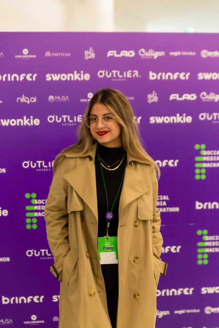 Mulher sorridente com óculos, cabelo castanho, vestindo casaco bege, brincos dourados e uma camiseta preta, usando crachá...