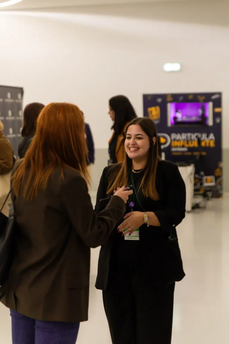 Mulher sorridente conversando com outra pessoa em evento, ao fundo cartaz de discussão inteligente.