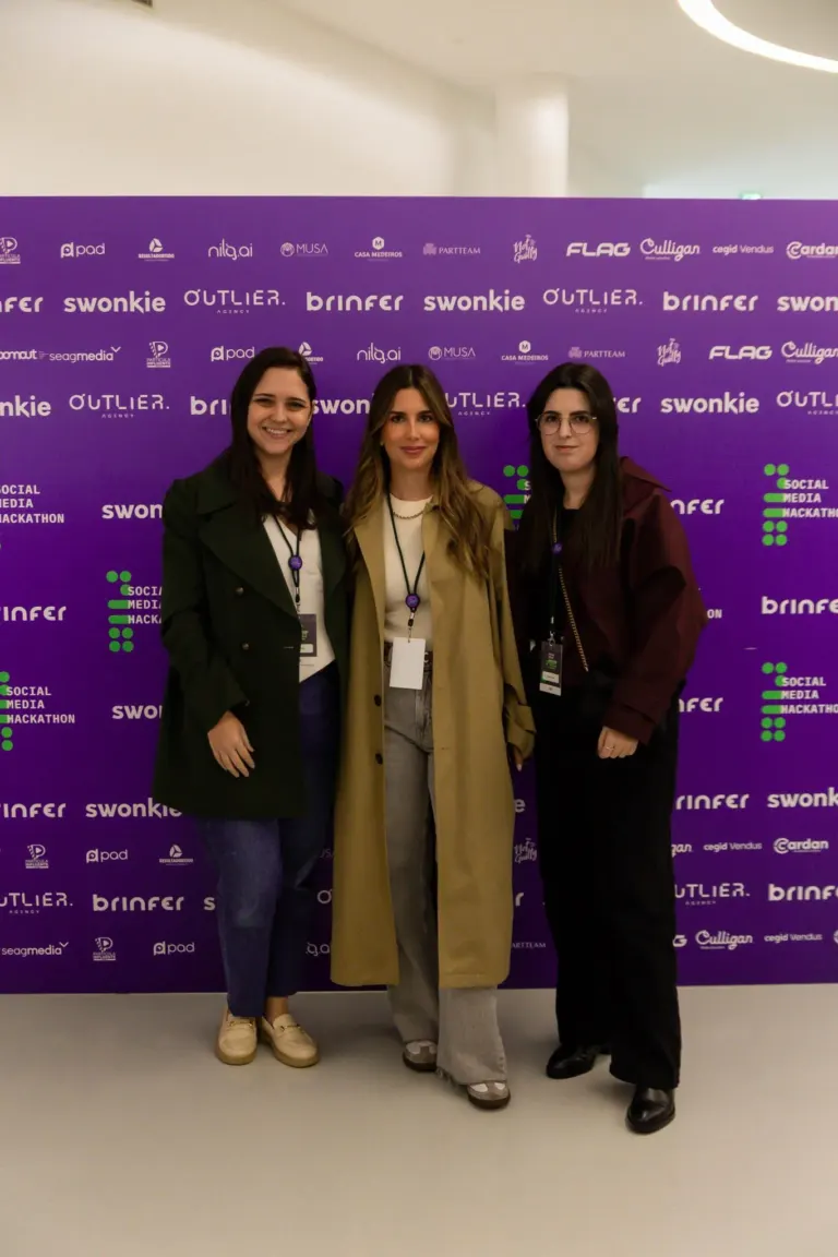 Três mulheres em pé na frente de um painel roxo com várias marcas e logotipos de eventos e empresas de mídia social.