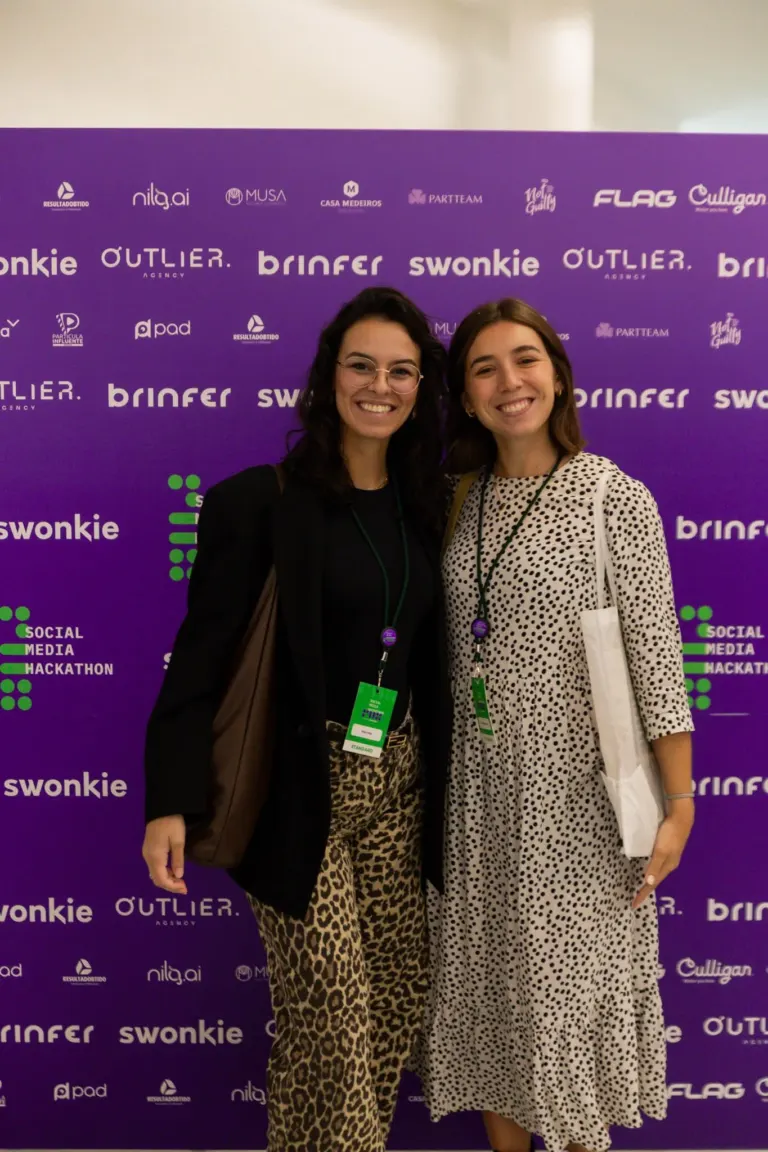Duas mulheres sorrindo posando para a câmera em evento com fundo roxo com logotipos de patrocinadores e inscrição de...