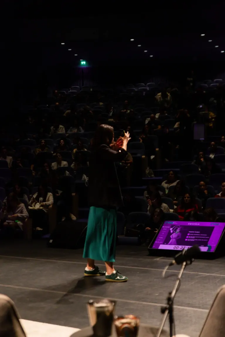 Pessoa no palco de um teatro, de costas, com roupas verdes e tênis, ao centro, com muitos espectadores sentados na...