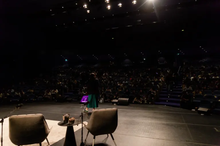 Cena de um teatro ou auditório com muitas pessoas sentado na plateia. No palco, há uma mulher com vestido verde, cabelos...