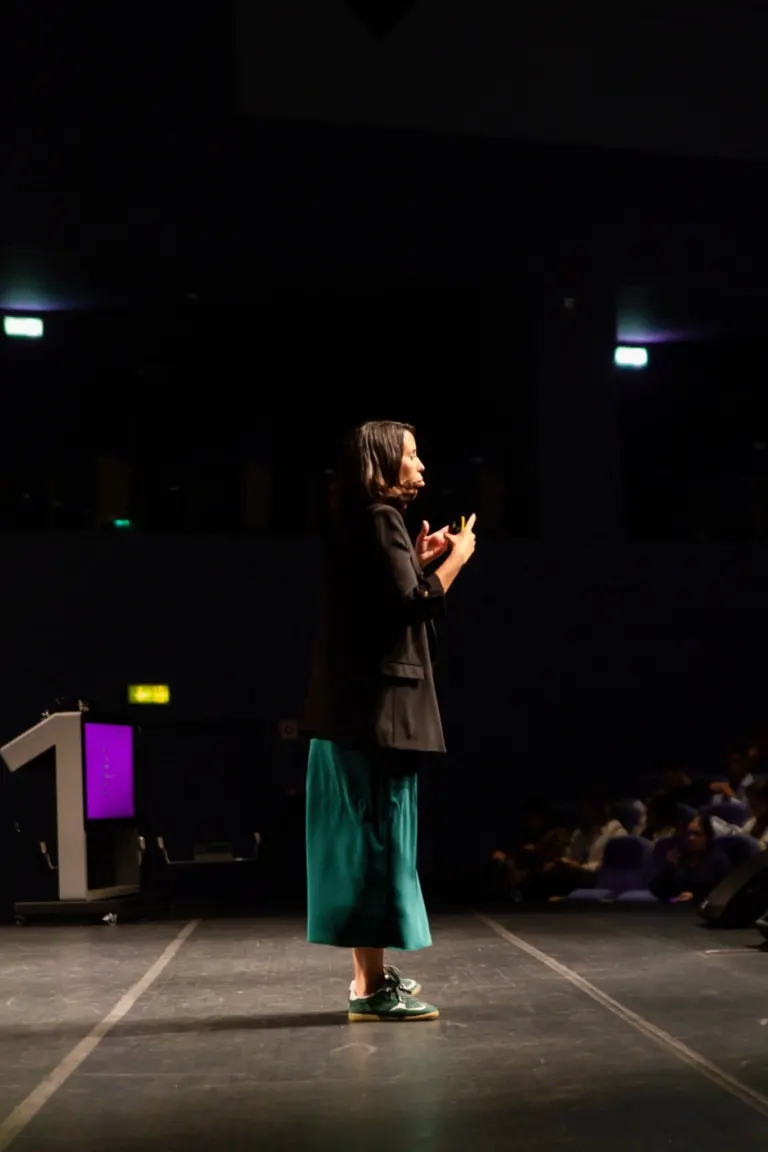 Mulher de cabelos castanhos atuando como palestrante em palco escuro com projeto de palco ao fundo, usando calça verde,...