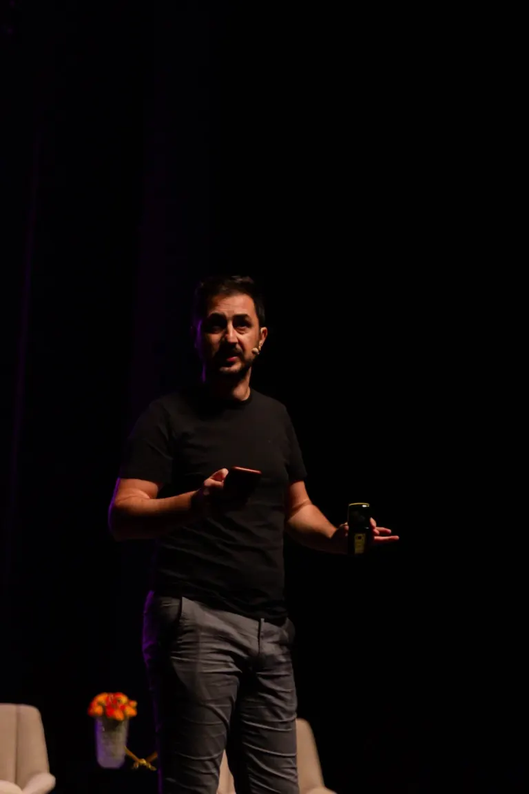 Homem com barba, usando uma camiseta preta e calça cinza, segurando um telefone e um microfone, em um palco com fundo...