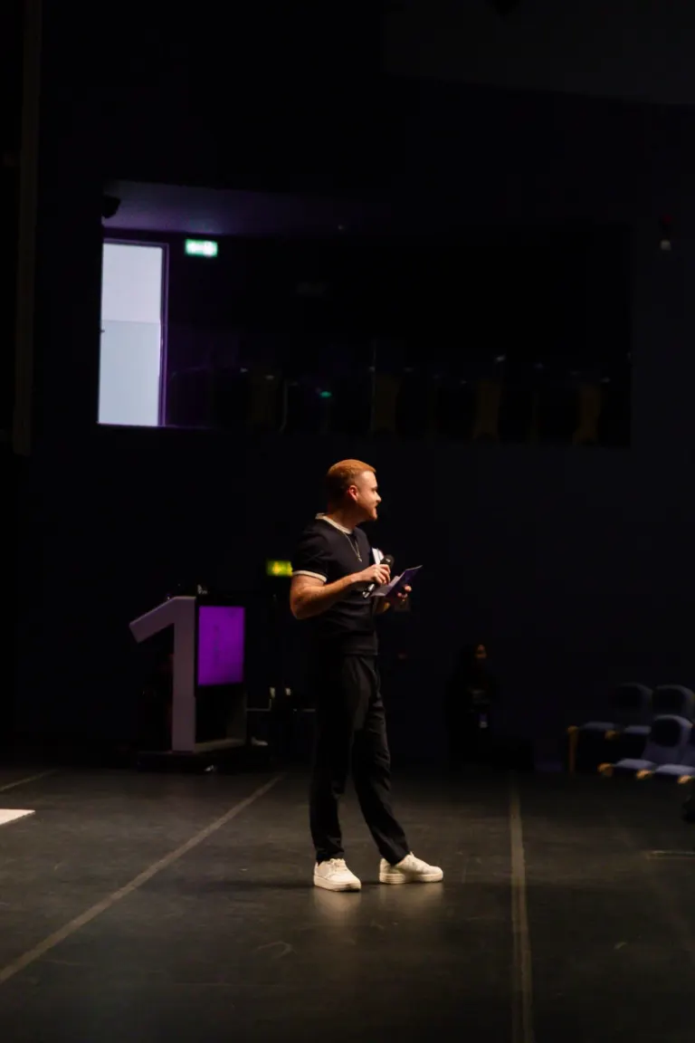 Pessoa em palco, segurando um microfone e um papel, com fundo escuro e assentos ao lado.