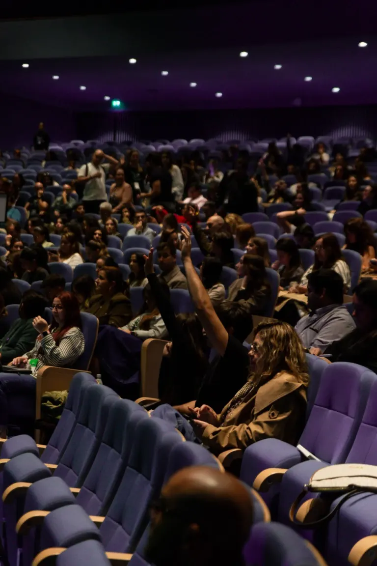 Público sentado em cadeiras de teatro ou cinema, algumas pessoas falando, outras usando smartphones, dentro de uma sala...