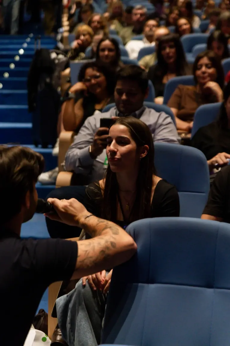Vários espectadores sentados em cadeiras azul escuro em uma sala de conferências, algumas pessoas sorrindo e usando...