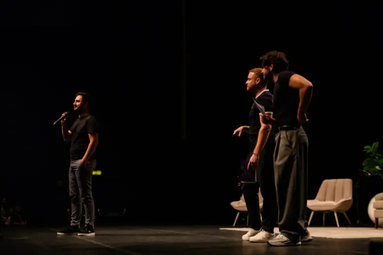 Homem com microfone falando em palco escuro, três homens observando, uma cadeira e sofá ao fundo.