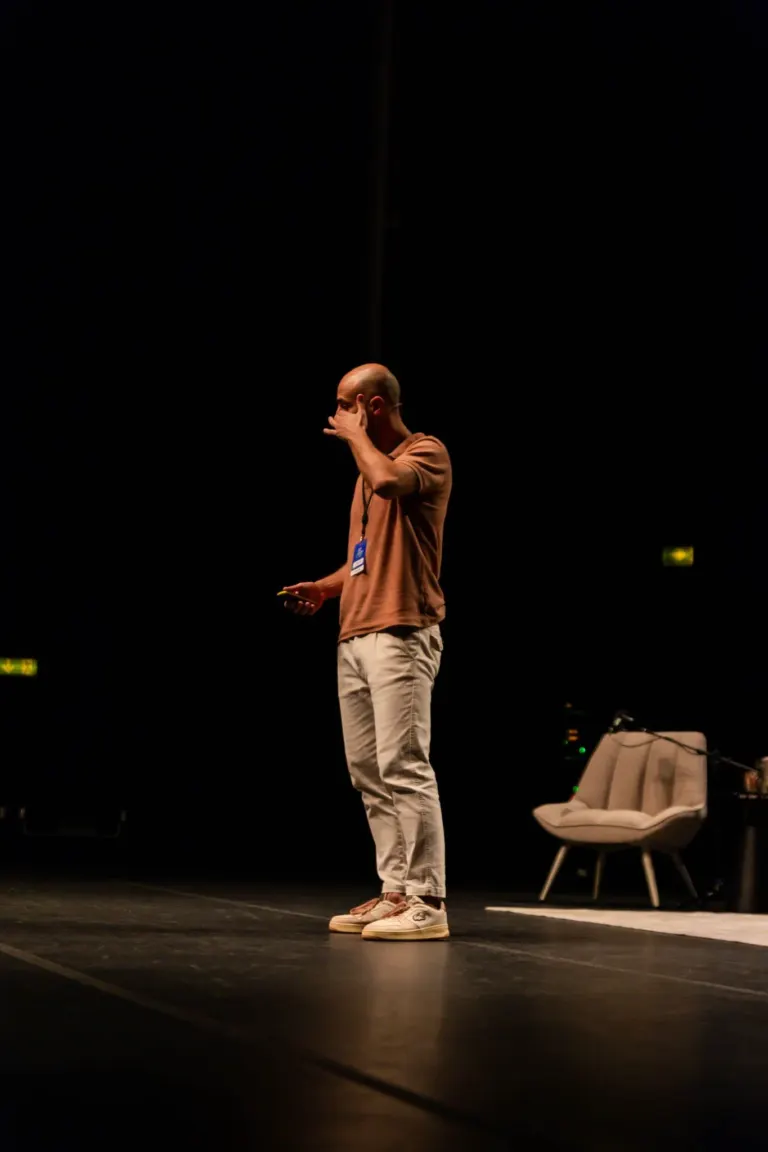 Homem de camisa laranja claro, calça bege e tênis branco, de cabeça raspada, segurando um celular, em pé no palco com...