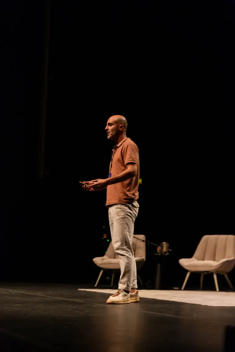 Homem de terno casual e tênis, segurando um celular, pensando durante uma apresentação em palco com cadeira e mesa ao fundo
