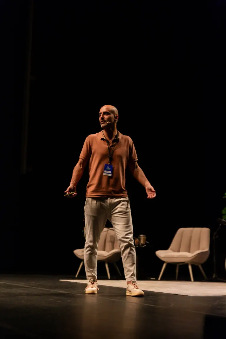 Homem com cabelo raspado, usando camiseta marrom, calça bege, tênis claro, com microfone em um palco com cadeiras ao fundo.