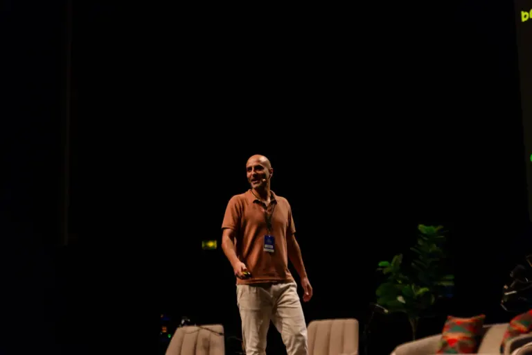 Homem de camiseta marrom e calça bege, falando na frente de um público, em um palco com fundo preto, segurando um...