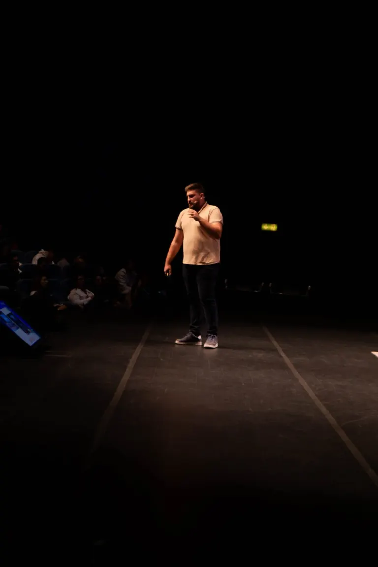 Homem de camiseta bege e calça jeans escura falando em palco em um evento com público sentado na plateia.