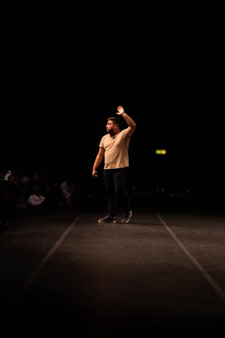 Homem de pé no palco, de camiseta bege e calça escura, segurando um dispositivo na mão esquerda, com o braço direito...