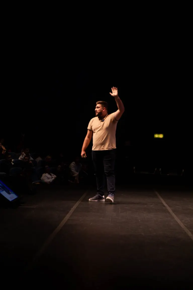 Homem de camiseta bege, calça cinza e tênis cinza, falando em palco com o braço levantado, em ambiente de apresentação ou...