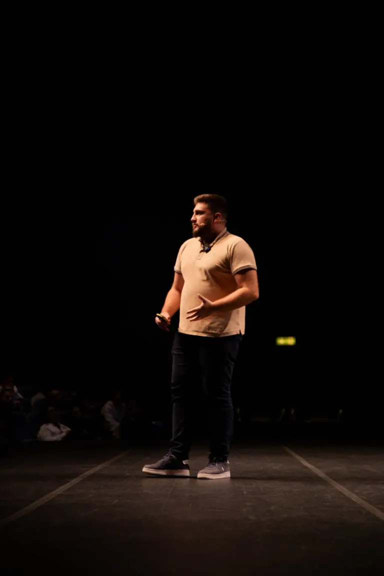 Homem de pé no palco, vestido com camiseta bege, calça escura e tênis cinza, falando com microfone no rosto, em um...