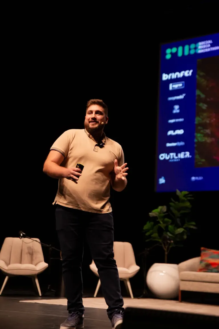 Homem com barba, usando uma camiseta bege, falando em um palco com um microfone de clipe e segurando um controle remoto,...