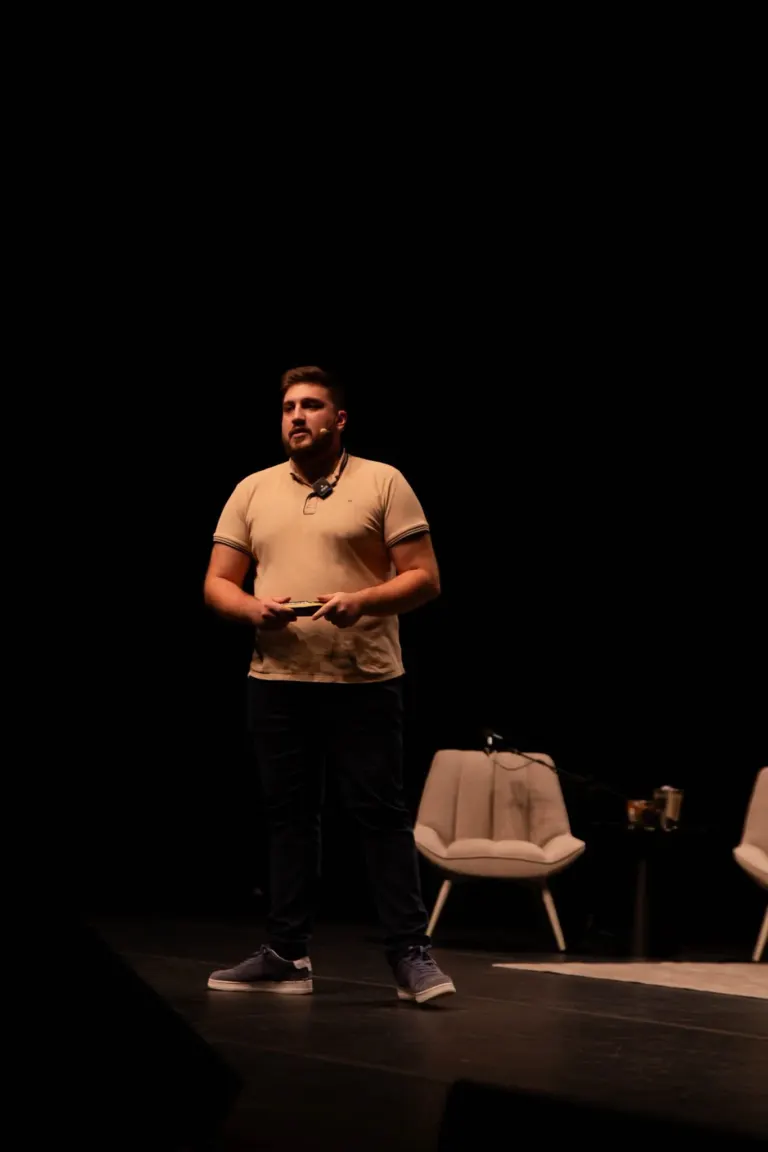 Homem em palco de teatro, de camiseta bege, com microfone de lapela, segurando um dispositivo, com cadeiras e mesa ao...