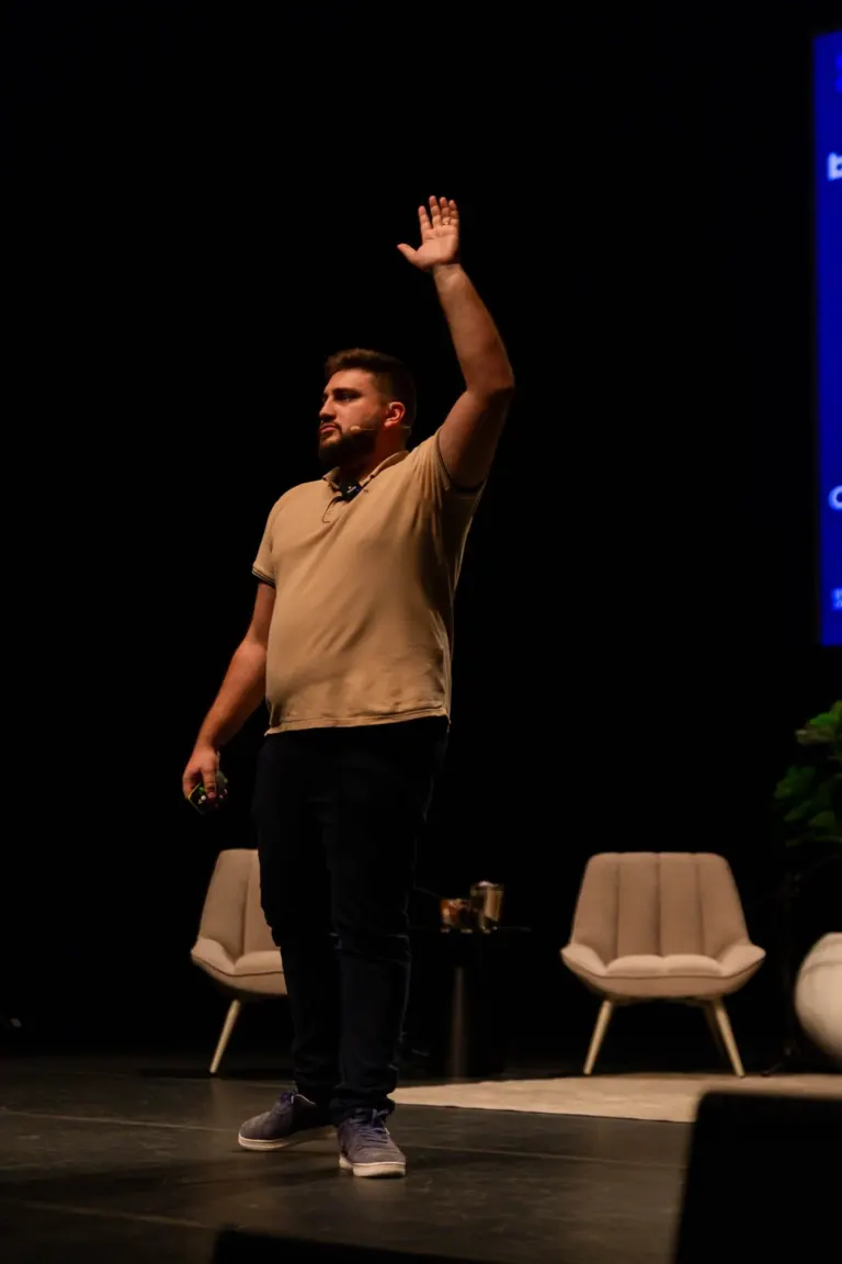 Homem em palco com braços levantados, sentado em cadeira com duas poltronas ao fundo.