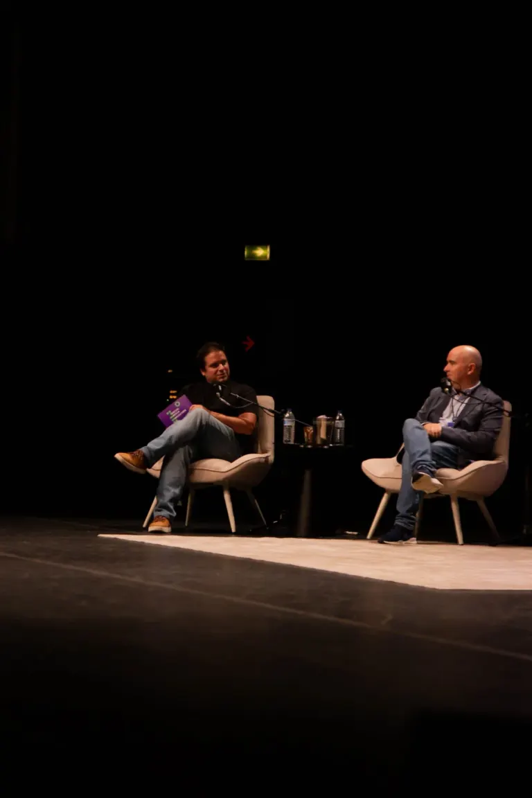 Dois homens participando de um painel de discussão em um palco de teatro, sentados em cadeiras com microfones, livros e...