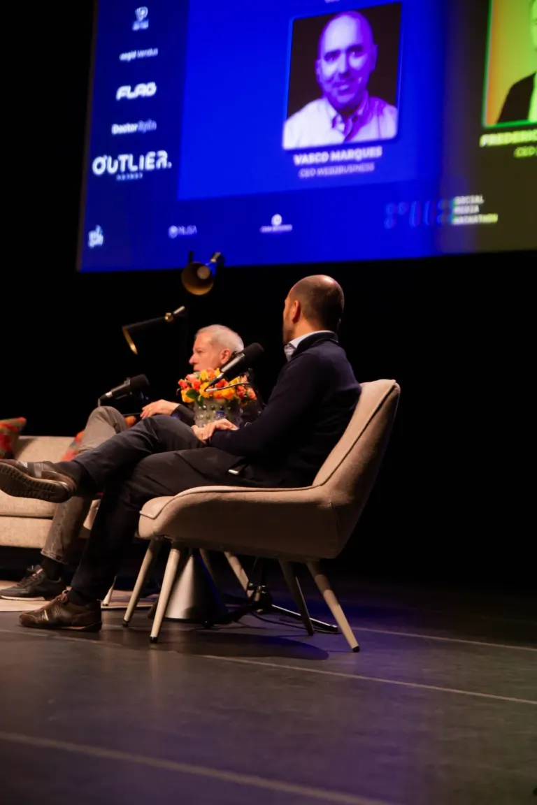 Imagem de uma apresentação ou painel de conferência com duas pessoas sentadas em cadeiras no palco. Um homem de cabelos...