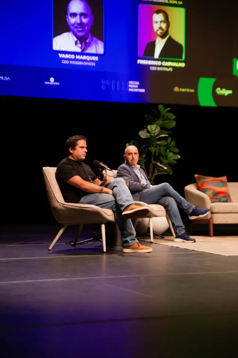 Imagem de dois homens sentados em cadeiras em uma sala de evento, com uma planta ao fundo, e uma tela grande acima deles...