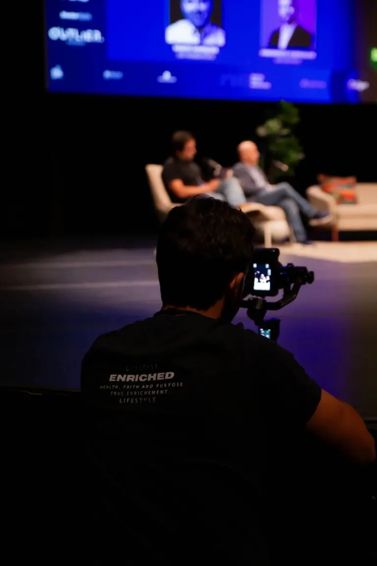 Pessoas participando de um painel ou discussão em um evento, com um público na plateia. Na parte inferior da imagem, uma...