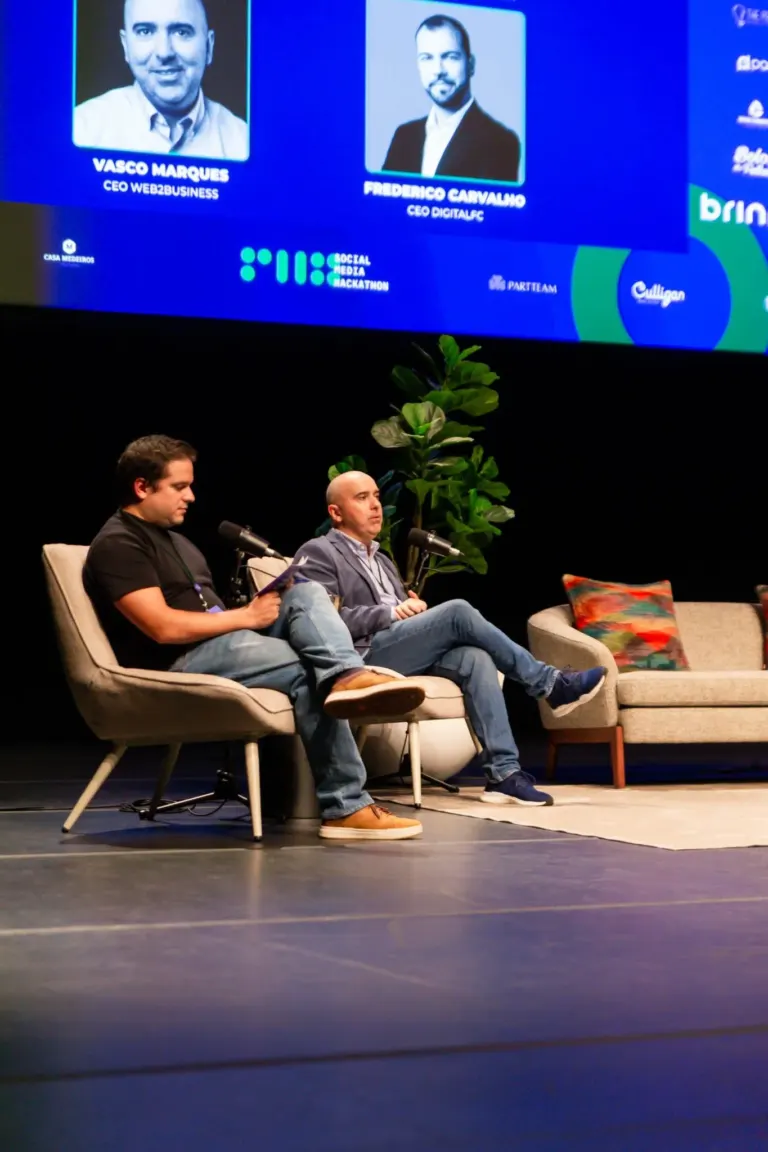 Imagem de um painel com fotos e nomes de empresários e uma mesa com dois homens sentados, um deles lendo um documento, em...