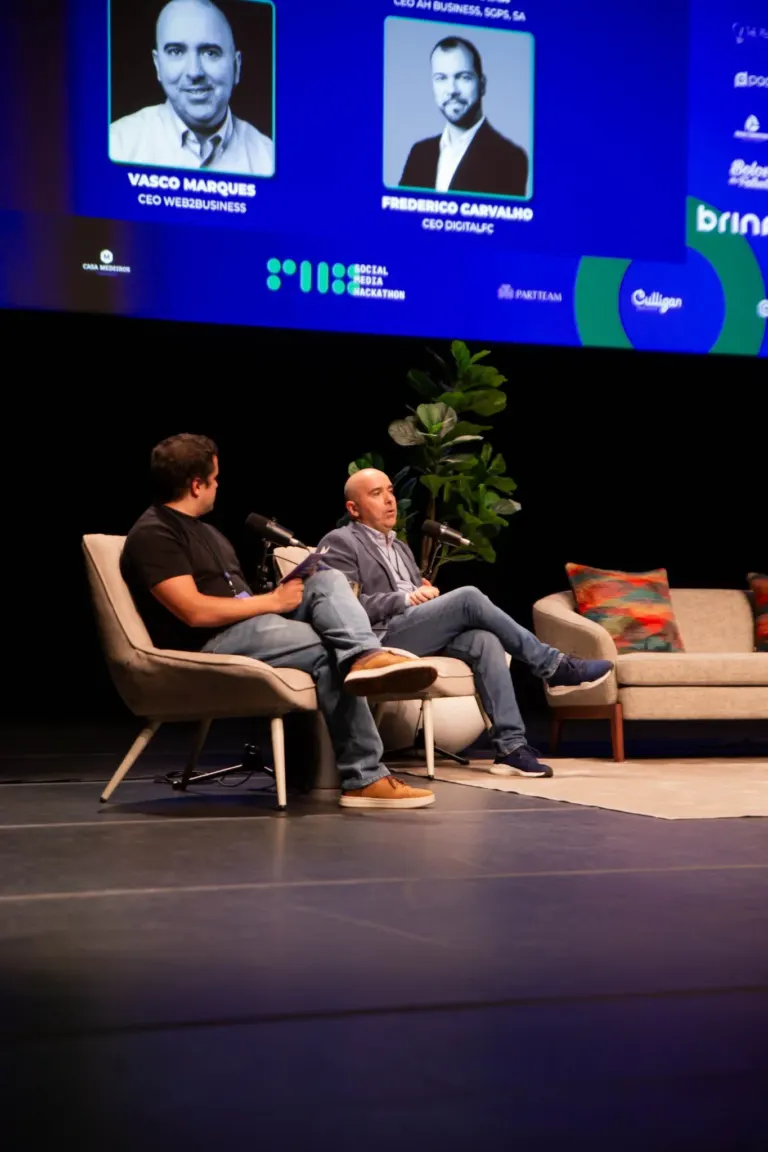 Duas pessoas sentadas em poltronas durante uma conferência, com um painel de fundo exibindo fotos e nomes de dois CEOs,...
