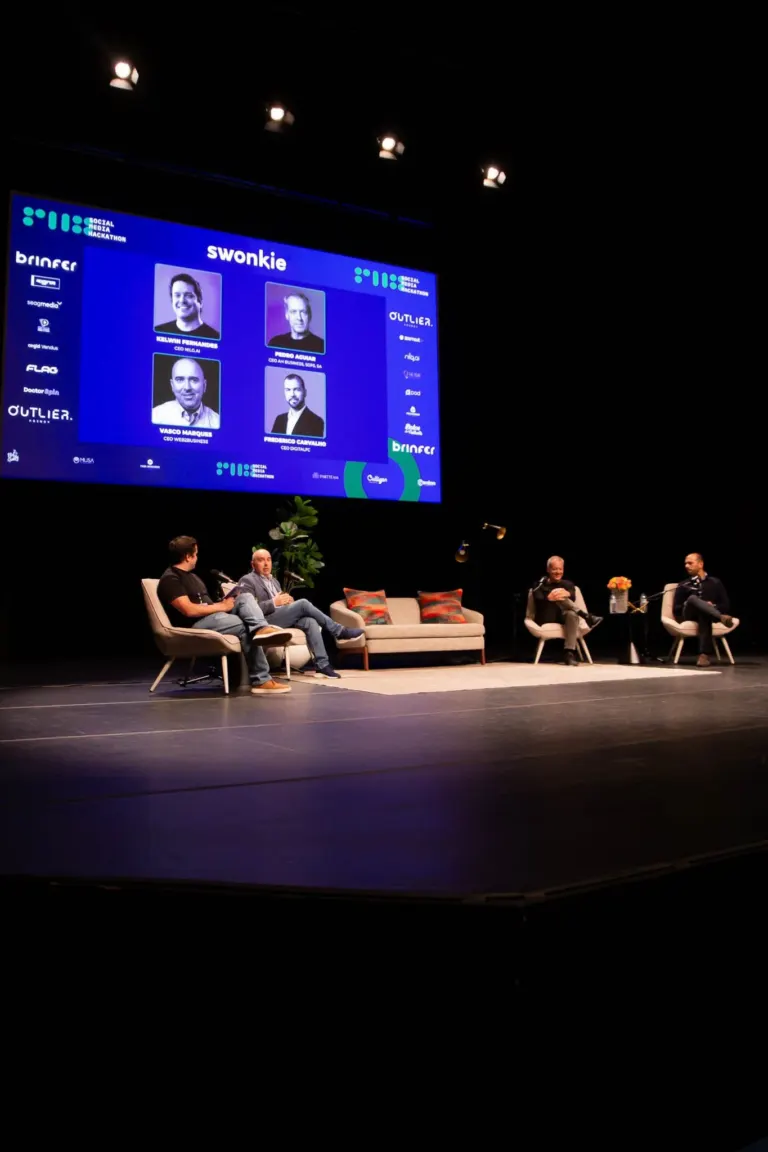 Imagem de uma apresentação ou painel de discussão em um palco com quatro pessoas sentadas em cadeiras, duas mulheres e...