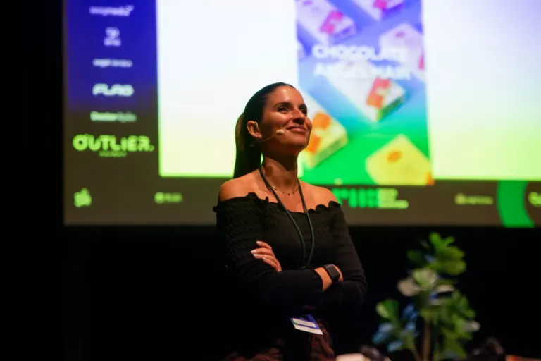 Mulher sorridente em um palco, usando um microfone de lapela, com uma tela de apresentação ao fundo que exibe imagens de...