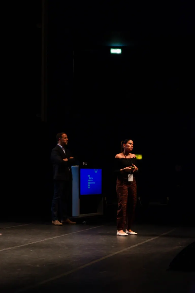 Pessoas em um palco, uma mulher fala ao microfone e um homem está ao lado num evento escuro com uma tela de computador ao...