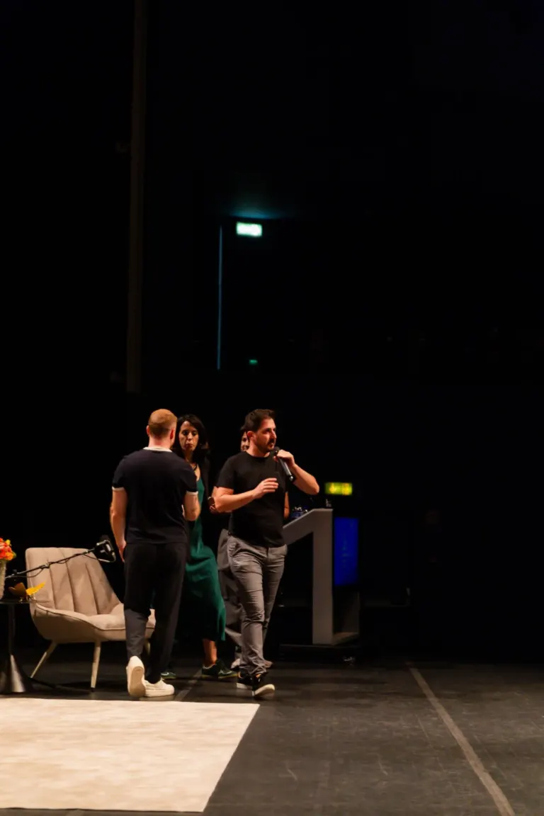 Quatro pessoas estão de pé em um palco de teatro, conversando. Há uma cadeira de aparência moderna e um arranjo de flores...