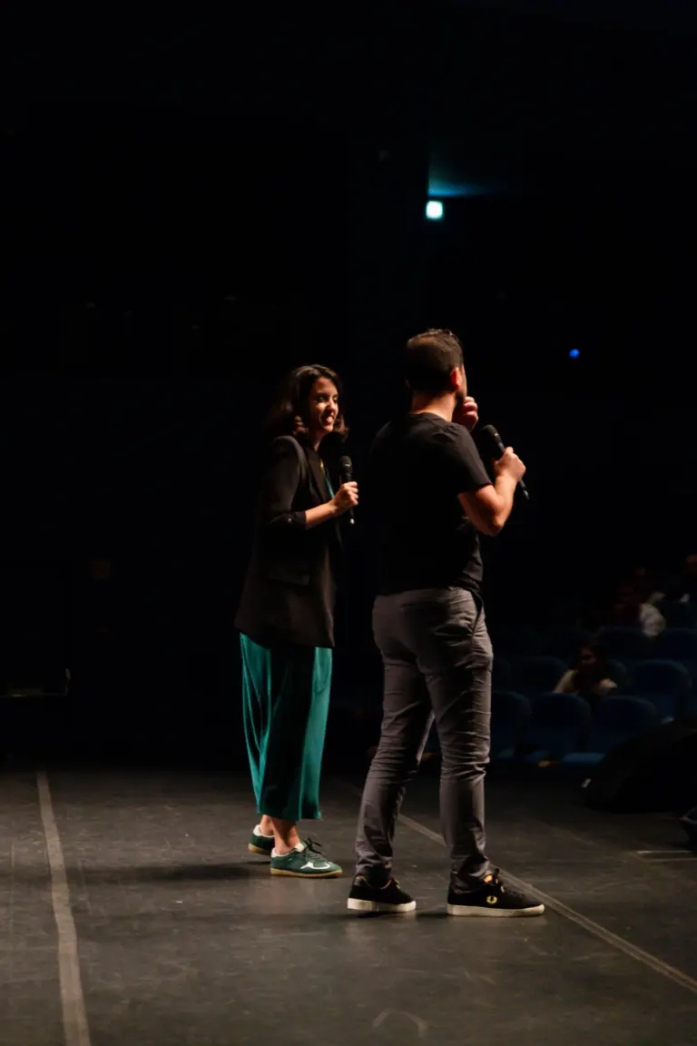 Duas pessoas no palco de um teatro, uma mulher e um homem, segurando microfones e conversando, com o palco vazio ao redor.