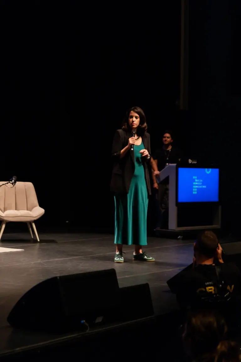 Mulher de cabelo castanho comprido, vestindo blusa azul, blazer preto e calça longa azul, segurando microfone em palco...