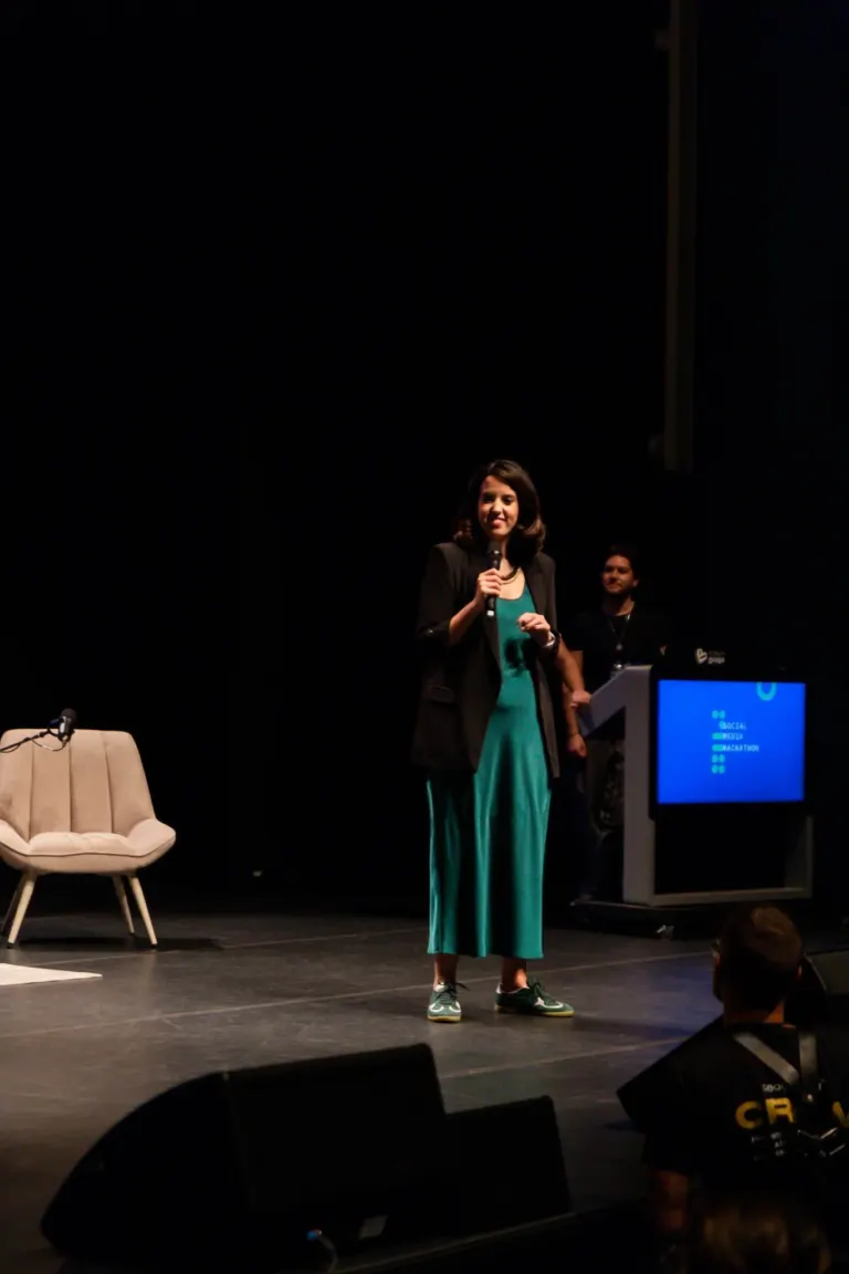 Mulher de cabelos castanhos ondulados, vestindo um vestido verde e um blazer preto, segurando um microfone, falando em um...