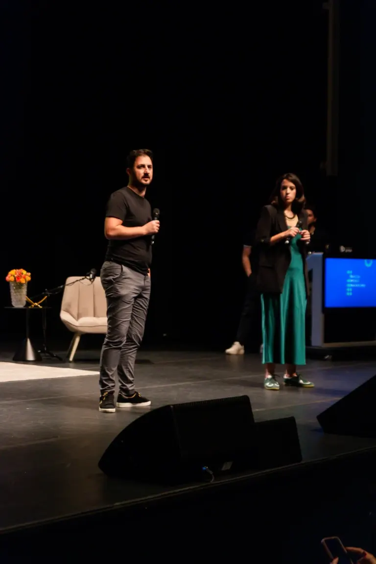 Homens e mulheres segurando microfones em um palco escuro, com cadeira, mesa com flores e tela exibindo apresentação ao...