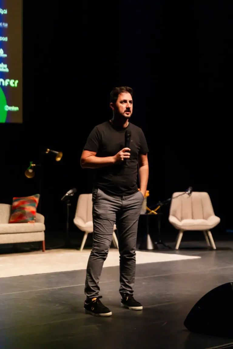 Homem segurando microfone em palco com cadeiras ao fundo, cenário de evento ou apresentação.