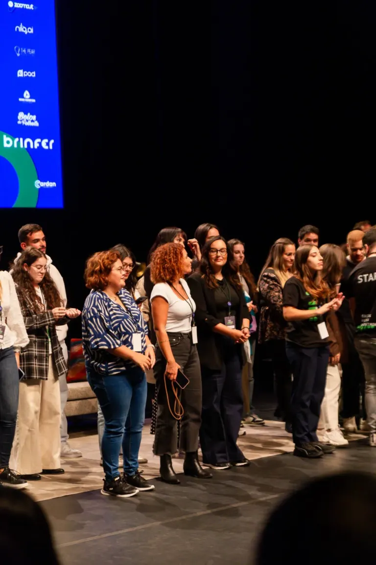 Grupo de pessoas em pé em fila, assistindo a uma apresentação ou evento em um palco, com uma tela grande ao fundo...