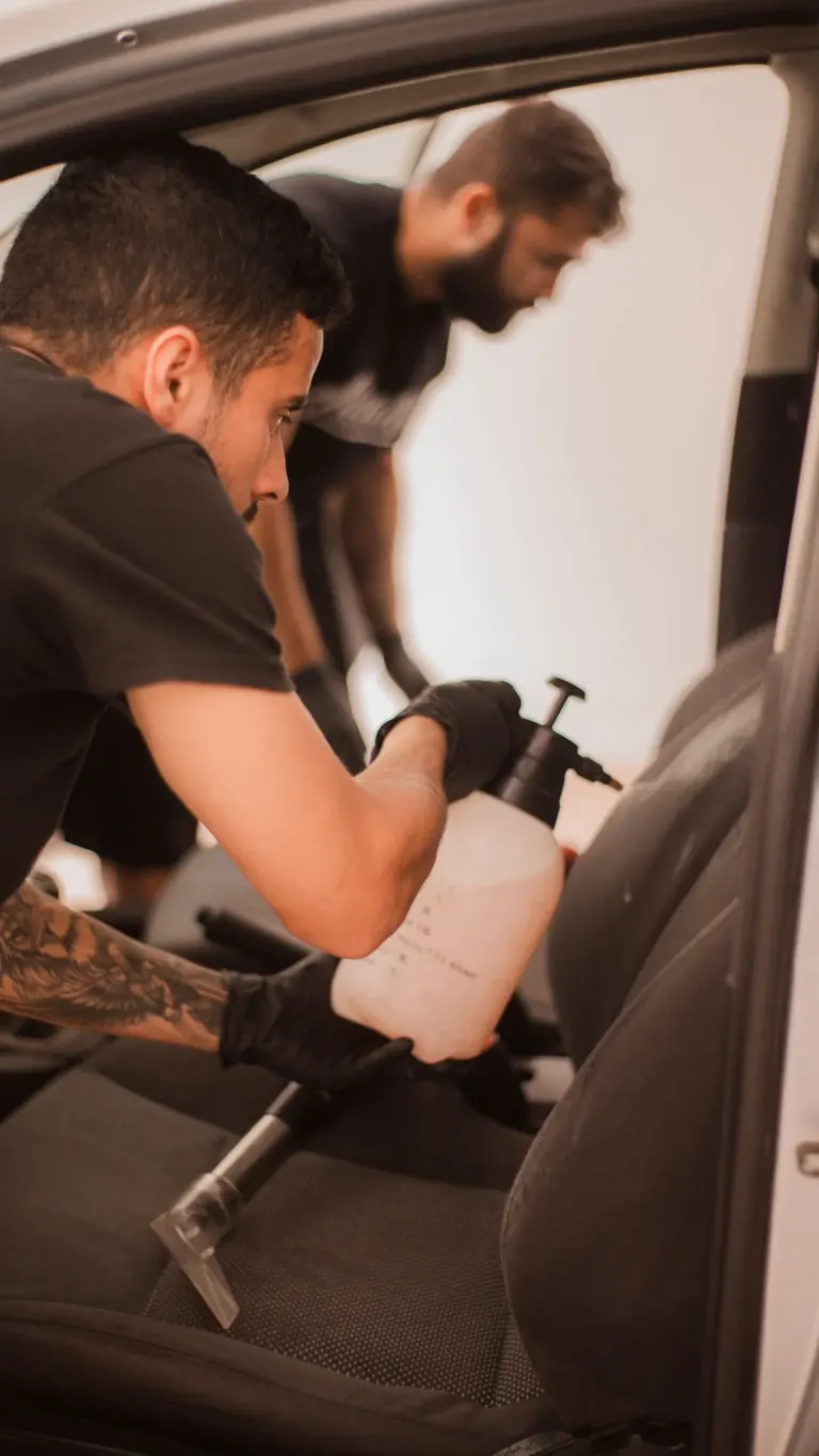 Dois homens limpando o interior de um carro, um deles segurando uma garrafa de spray e uma ferramenta de limpeza.