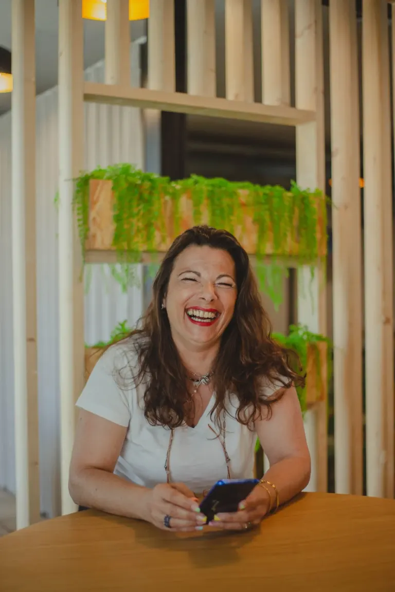 Mulher sorrindo, sentada à mesa, segurando um smartphone, com decoração de plantas verdes ao fundo.